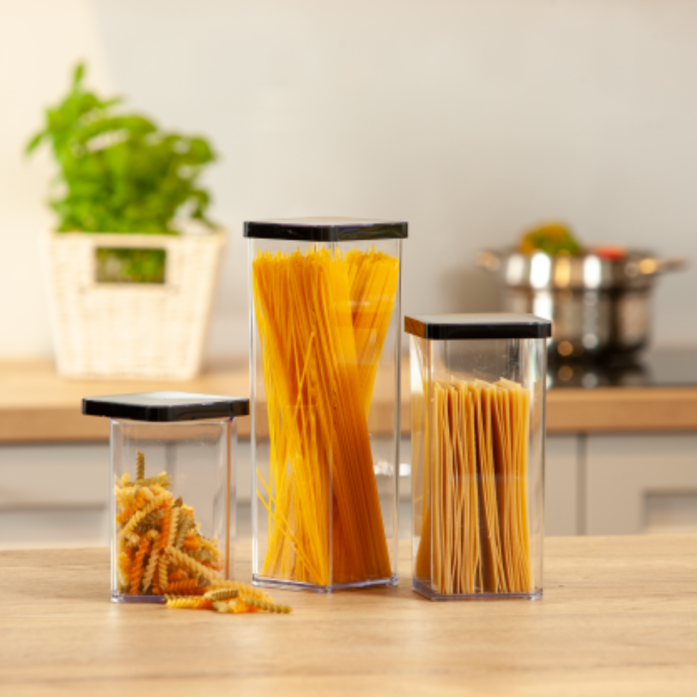 Drei Rotho Vorratsdosen-Set LOFT schwarz (6-teilig) mit schwarzen Deckeln halten Fusilli, Spaghetti und Linguine auf einer hölzernen Küchenzeile. Unscharf im Hintergrund stehen eine Pflanze und ein Topf - ideal für die Vorratsschrank Organisation.