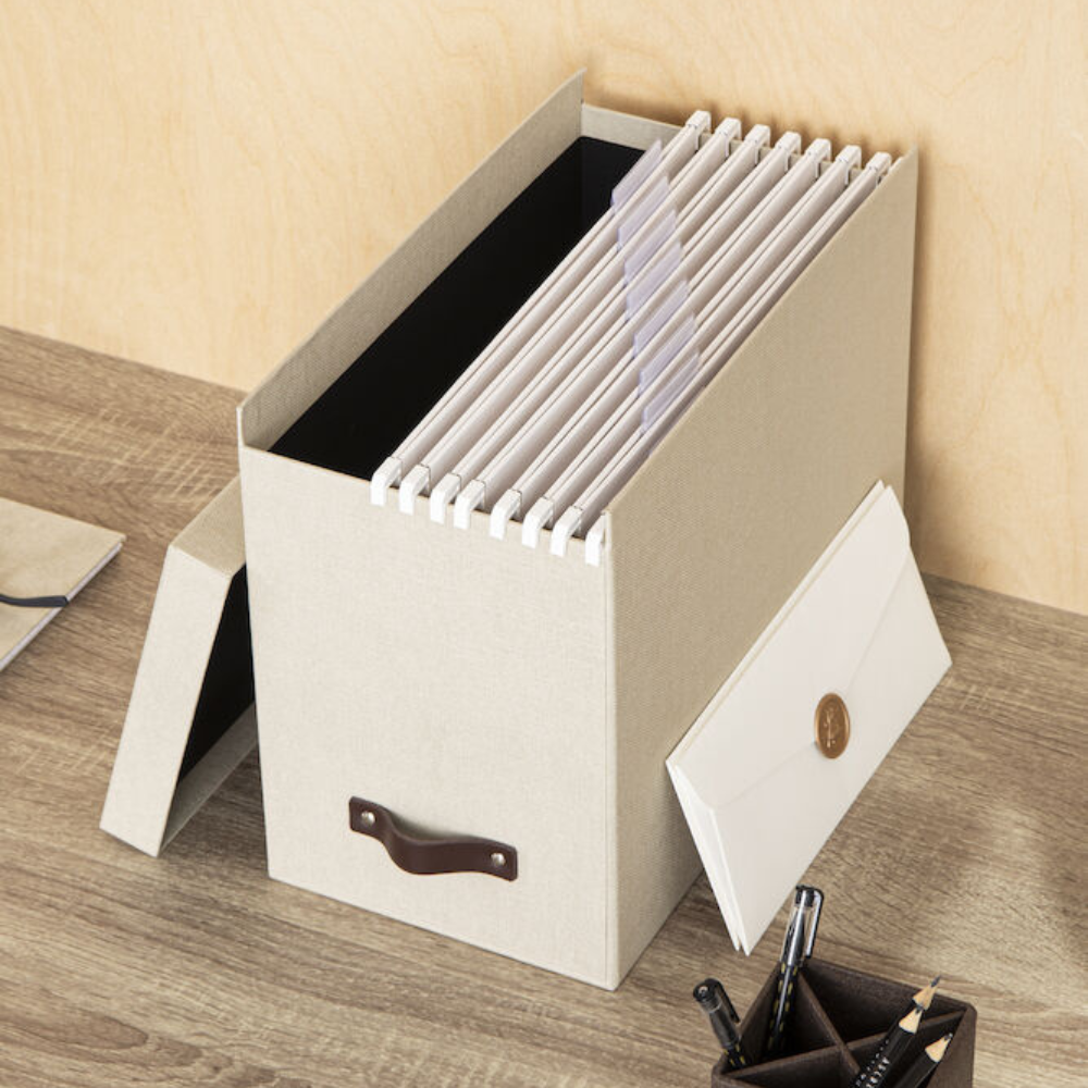 Die Bigso JOHAN Hängeregister-Box aus Linnen Canvas mit braunem Griff steht auf einem Holzschreibtisch und fasst A4-Ordner und einen Umschlag. In der Nähe befinden sich Schreibwaren für die einfache Organisation von Dokumenten.