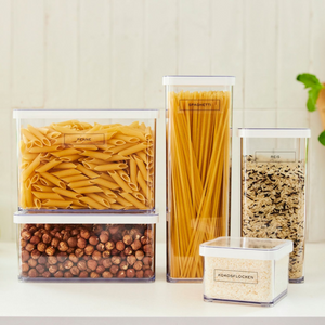 Fünf durchsichtige Plastikbehälter mit Penne, Spaghetti, Reis, Haselnüssen und Kokosflocken stehen fein säuberlich auf einer hellen Küchentheke, jeder mit Rotho's LOFT Aufkleber - Küchenorganisation zur einfachen Identifizierung beschriftet.