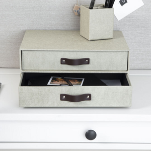 Auf einem weißen Schreibtisch steht die Bigso BIRGER Schubladenbox 2 Fächer aus Leinen Canvas in Hellgrau, die untere Lade offen mit Papieren und Foto; daneben ein passender Stiftehalter vor heller Wand.