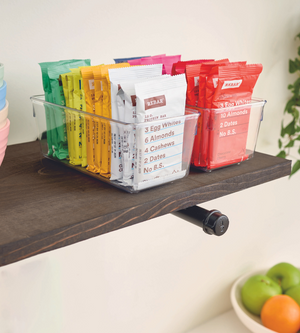 Ein iDesign LINUS - Schubladenorganizer HOCH hält bunte RXBAR-Proteinriegel auf einem dunklen Holzregal, darunter ist eine Schale mit Obst zu sehen.