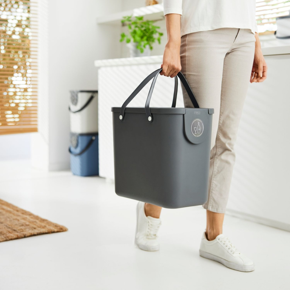 Eine Person in heller Hose und weißen Turnschuhen trägt einen großen anthrazitfarbenen ALBULA Recycling Müllsystem 25L von Rotho mit Griffen in einem hellen, modernen Raum, der mit Pflanzen und natürlichem Licht gefüllt ist.