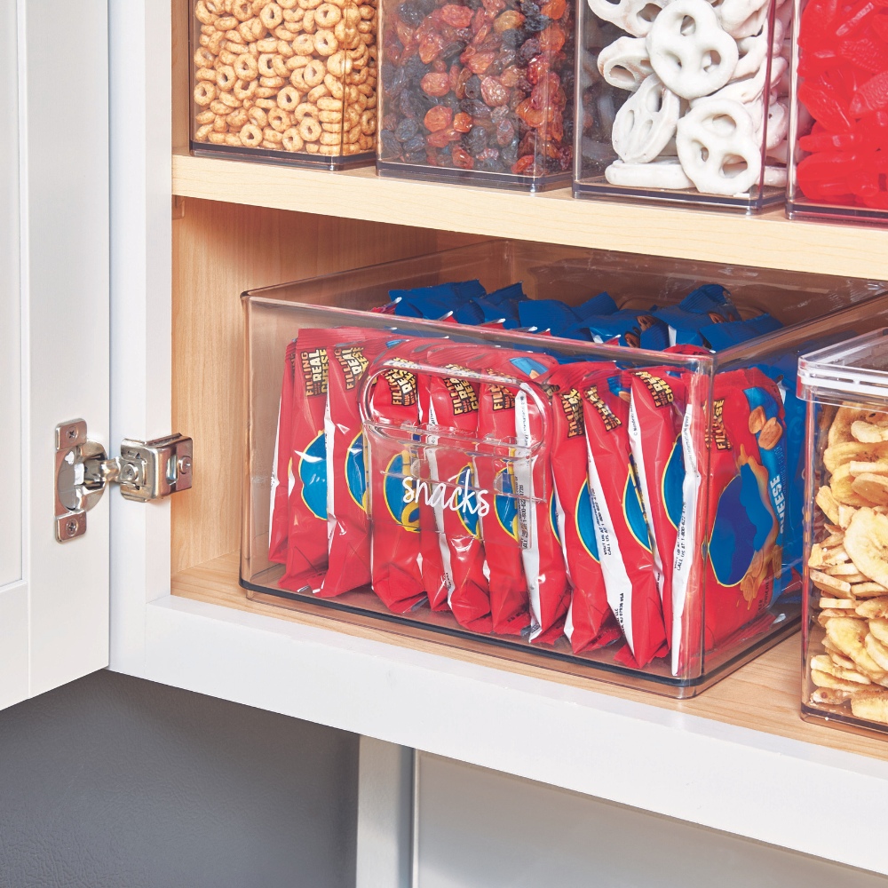 Ein durchsichtiger iDesign HOME EDIT Etikettenhalter KLAR mit der Aufschrift "Snacks" enthält rote Snacktüten in einer Speisekammer, umgeben von anderen beschriftbaren Behältern mit Müsli, Trockenobst, Brezeln und Chips.