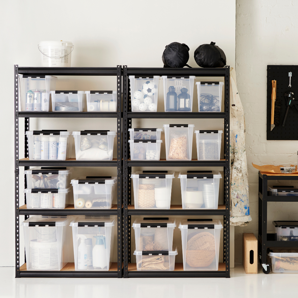 Zwei schwarze Metallregale enthalten durchsichtige Orthex SmartStore Box KLAR 31L, die BPA-frei und stapelbar sind und Haushaltsgegenstände enthalten. In der Nähe stehen ein Gerätewagen, eine Hängeschürze, ein Eimer und eine Stecktafel mit Werkzeugen vor einer weißen Wand.