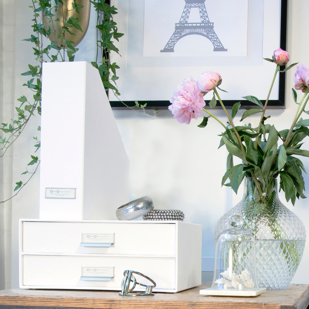 Ein aufgeräumter Schreibtisch mit weißen Schubladen, die Bigso VIKTORIA Stehsammler Büroablage - Weiss mit Etikettenhalter, eine Glasvase mit rosa Pfingstrosen, eine Glascloche mit weißer Koralle, grüne Ranken und ein gerahmter Eiffelturm-Druck an der Wand.