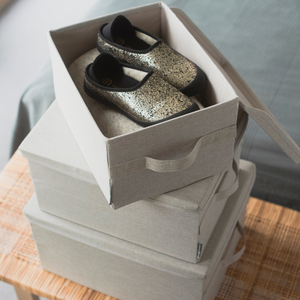 Ein Paar schwarz-goldene Krabbelschuhe sitzt in einer offenen, zusammenklappbaren Box auf zwei geschlossenen, stapelbaren SOFT Aufbewahrungsbox Beige L - 45x34x25cm von Bigso auf einer Holzfläche neben einem Bett.