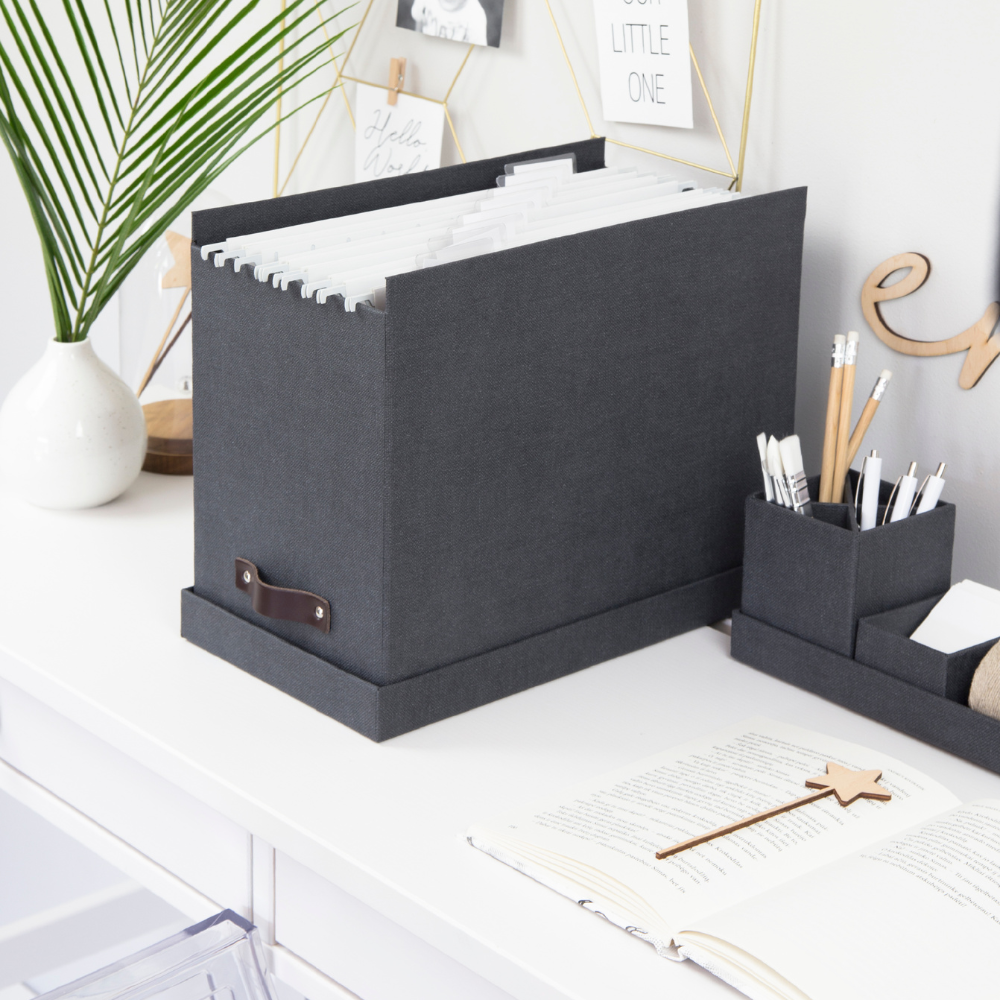 Graue Ablagebox mit Hängeordnern auf einem weißen Schreibtisch, umgeben von Dekorationen.