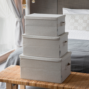 Drei Bigso SOFT Aufbewahrungsboxen in Beige (Größe M, 40x30x22 cm) sind im Schlafzimmer auf einer Bank gestapelt; im Hintergrund sieht man ein ordentlich gemachtes Bett und ein Fenster mit transparenten Vorhängen.