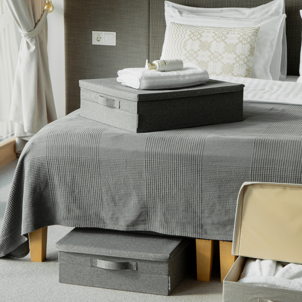 Ein ordentlich gemachtes Bett mit einer grauen, strukturierten Tagesdecke und Kissen verfügt über eine SOFT Aufbewahrungsbox Grau - Unterbett-Lagerung von Bigso für intelligenten Stauraum unter dem Bett, während ein offener Koffer am Fußende des Bettes steht.