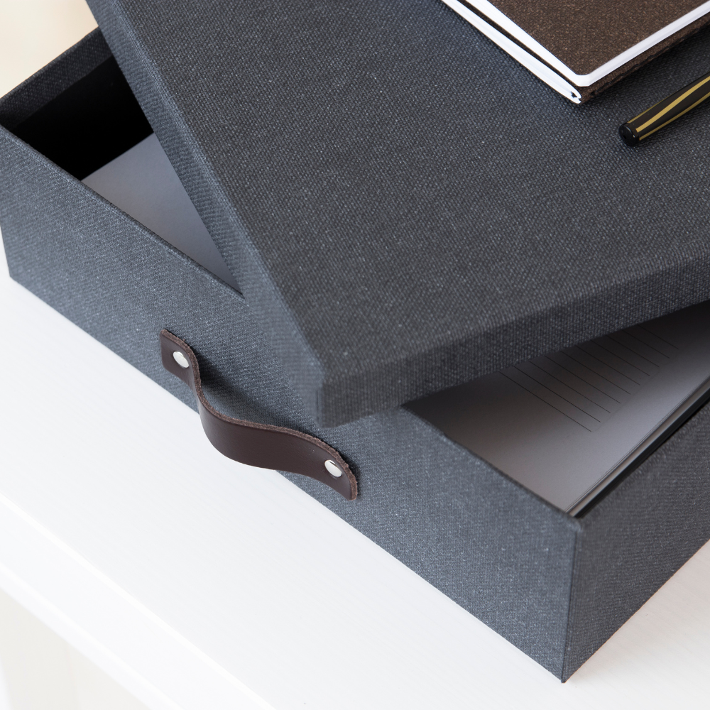 Eine Bigso OSKAR Aufbewahrungsbox A4 in Schwarz Canvas mit einem Ledergriff liegt leicht geöffnet auf einer weißen Fläche und zeigt ein Notizbuch im Inneren. Ein weiteres Notizbuch und ein Stift liegen oben auf der stilvollen Aufbewahrungsbox.