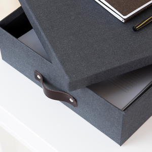 Eine Bigso OSKAR Aufbewahrungsbox A4 in Schwarz Canvas mit einem Ledergriff liegt leicht geöffnet auf einer weißen Fläche und zeigt ein Notizbuch im Inneren. Ein weiteres Notizbuch und ein Stift liegen oben auf der stilvollen Aufbewahrungsbox.