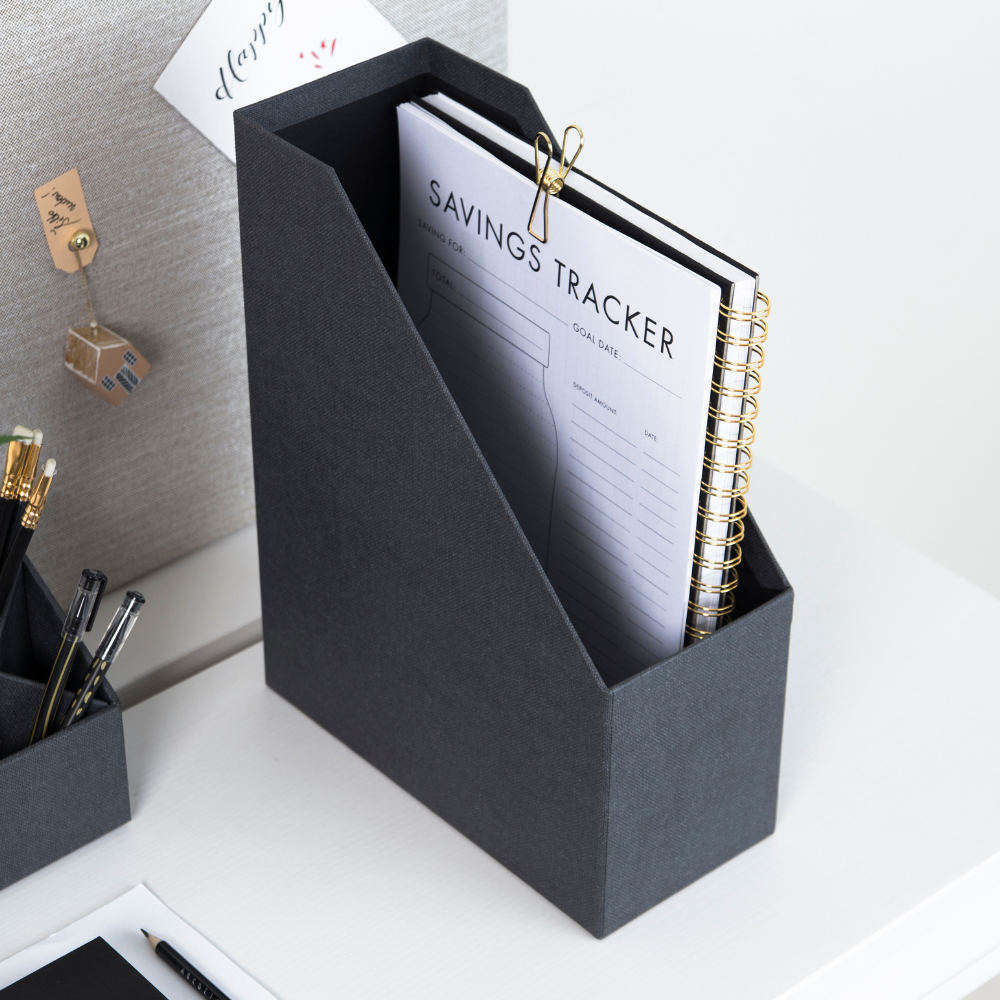 Die schwarze ESTELLE Stehsammler Büroablage von Bigso aus Canvas steht auf einem weißen Schreibtisch, hält ein mit Gold-Clip befestigtes "Savings Tracker"-Spiralnotizbuch sowie Stifte und Bürobedarf und sorgt für ordentliche A4-Aufbewahrung.