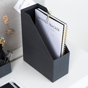 Die schwarze ESTELLE Stehsammler Büroablage von Bigso aus Canvas steht auf einem weißen Schreibtisch, hält ein mit Gold-Clip befestigtes "Savings Tracker"-Spiralnotizbuch sowie Stifte und Bürobedarf und sorgt für ordentliche A4-Aufbewahrung.